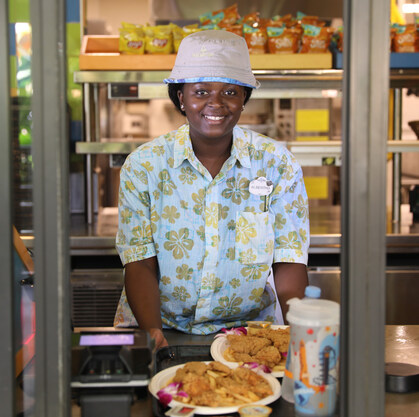 A woman is standing in a window smiling, at her hands are two plates of food that she is giving to a guest,
