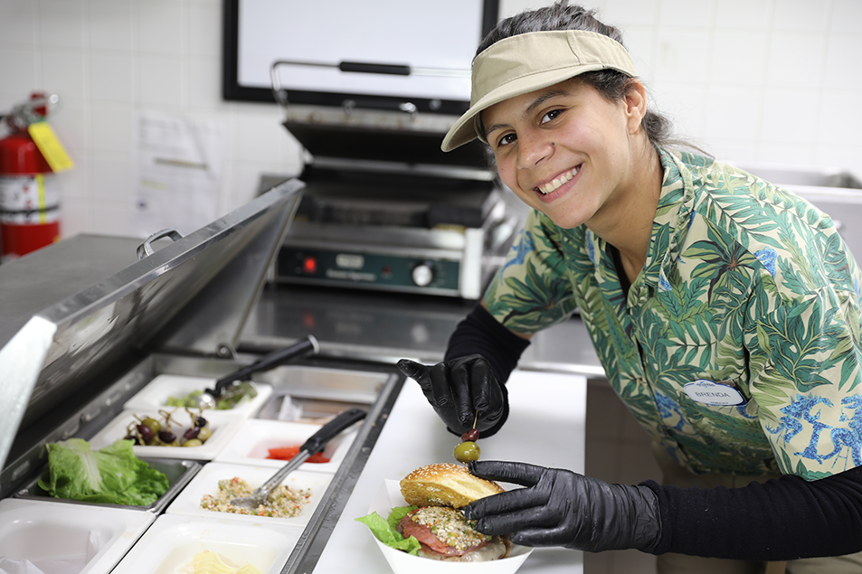 Woman is smiling and standing in a kitchen is prepping a burger.