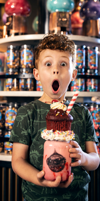 Child Looking into the camera while holding a pink milkshake that is topped with a slice of cake.
