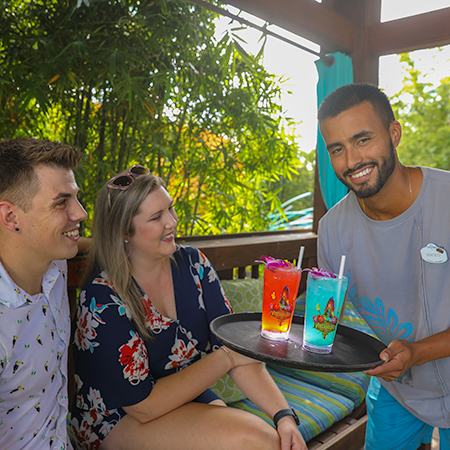 A couple is sitting together, smiling up at their waiter who is holding a tray with two drinks.