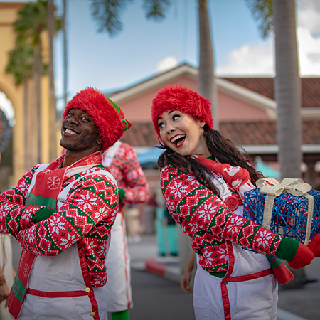 Two holiday performers dressed in red stand back-to-back and look back at each other. The performer on the right is holding a blue present.