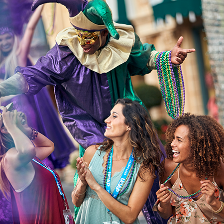 A Mardi Gras performer on stilts dressed as a jester gives out bead necklaces to a group of guests.