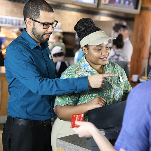 A Food and Beverage Manager assists a Team Member complete a transaction at the cash register.