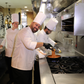 two chefs in white coats preparing pasta in a kitchen.