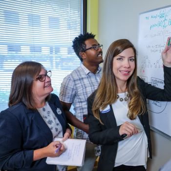 a woman in a black blazer stands next to a white board in an office setting, pointing at plans while working with two individuals.