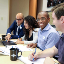 A team of professionals gathers around a table, utilizing a laptop to share ideas and enhance their project.