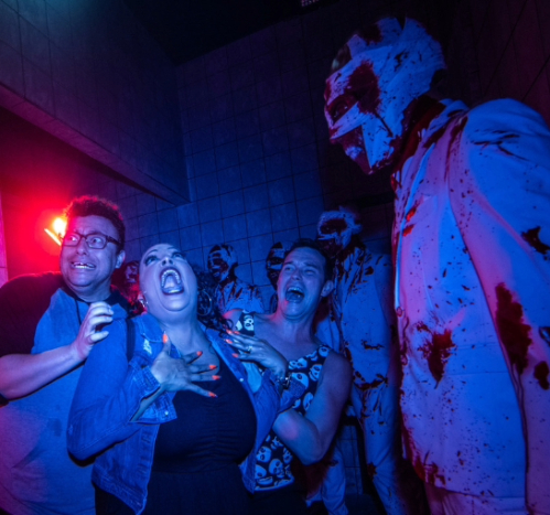 A Halloween performer in mask scares woman walking through haunted house, who appears frightened with her mouth open.